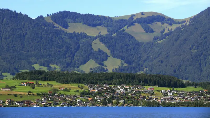 Dorf am Seeufer mit bewaldeten Hügeln im Hintergrund